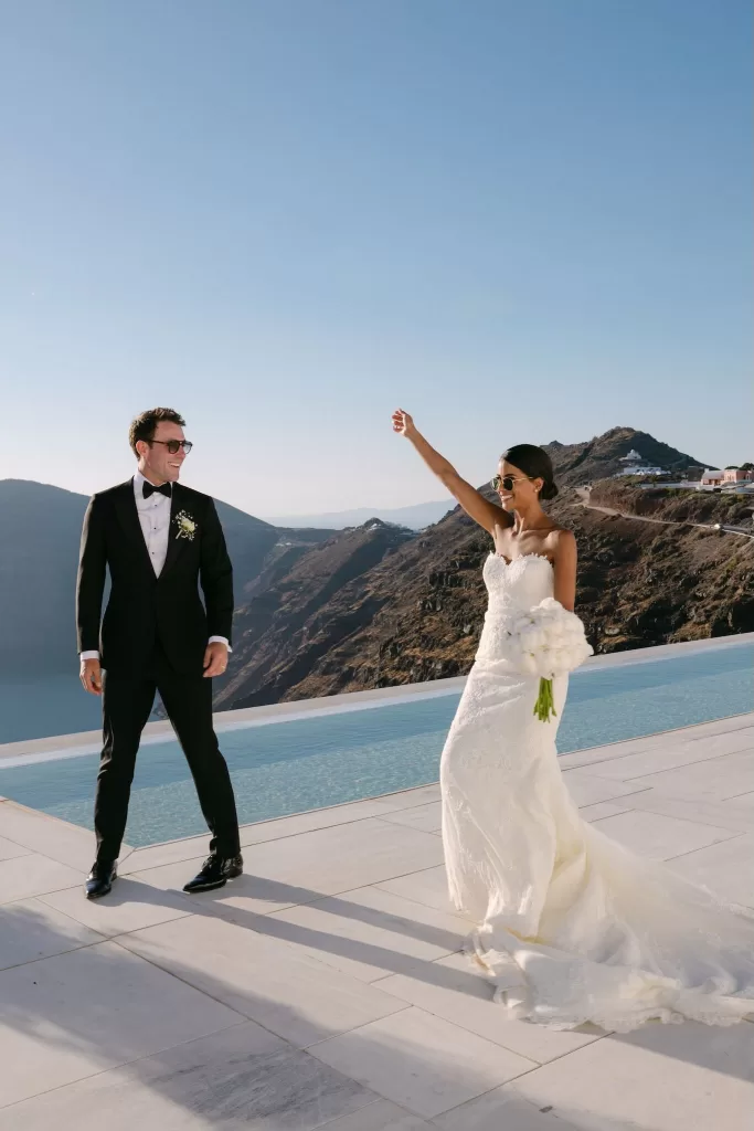 Joyful bride throws her arms up in celebration with Santorini’s whitewashed cliffs behind her