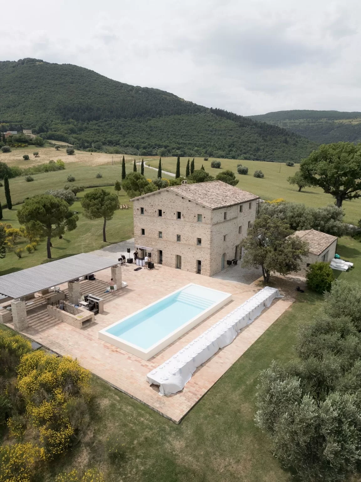 Aerial view of Casa Olivi surrounded by rolling hills, with a long poolside dining setup and rustic stone house.