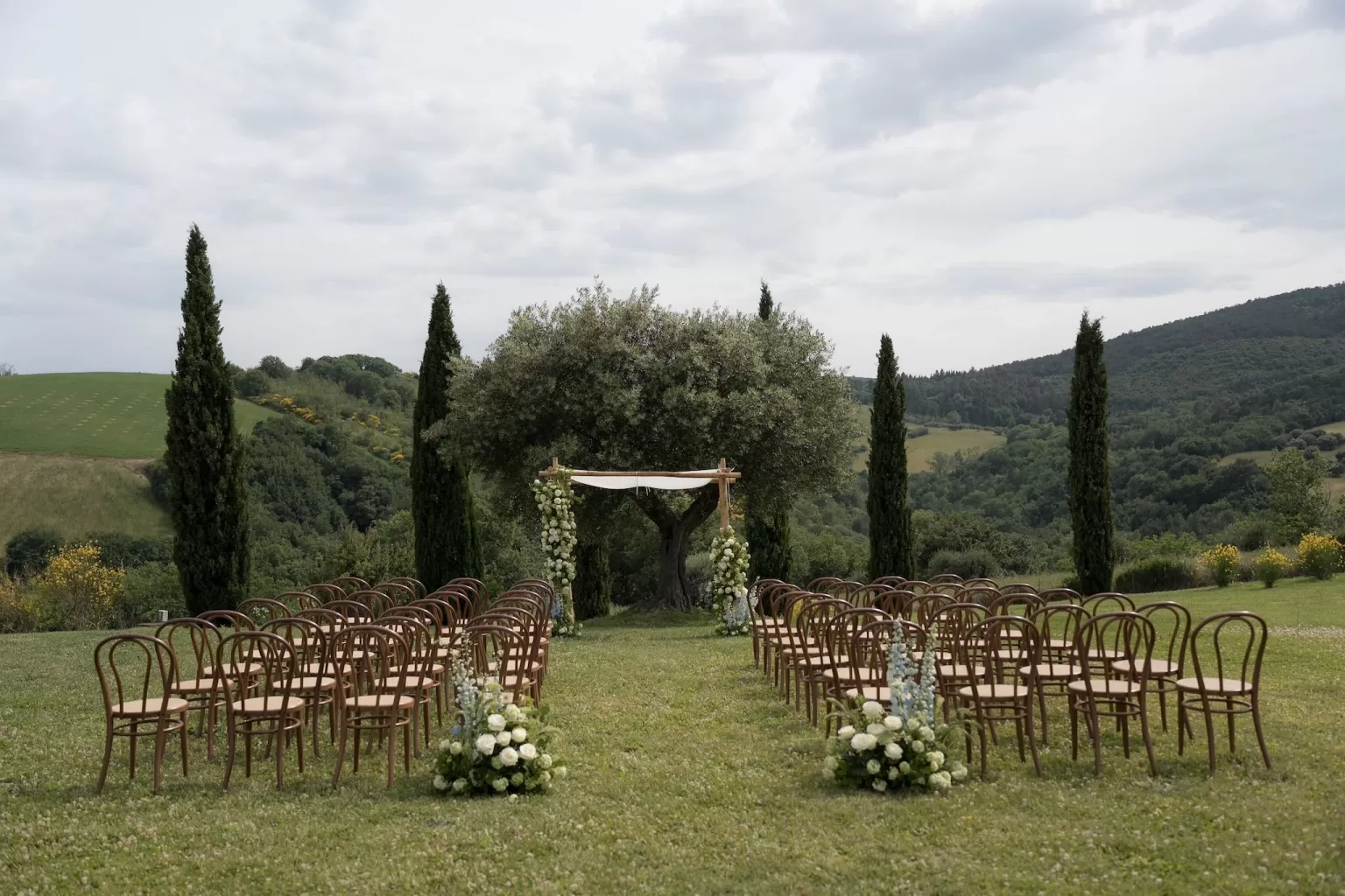 Outdoor wedding ceremony setup at Casa Olivi with a floral arch and wooden chairs overlooking green hills.