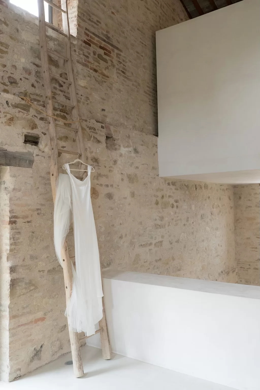Minimalist wedding dress and veil hanging on a wooden ladder against the rustic stone interior of Casa Olivi.