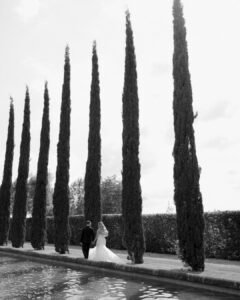 The couple stands between tall cypress trees, their romantic embrace framed by the Tuscan-inspired landscape of Domaine d'Essendieras.