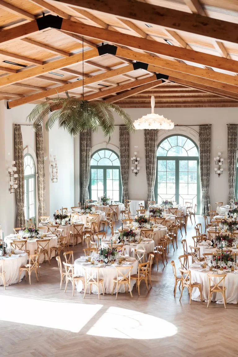 Wide view of the wedding reception set up inside Finca La Concepción’s grand hall, with wooden ceilings, arched windows and round tables.