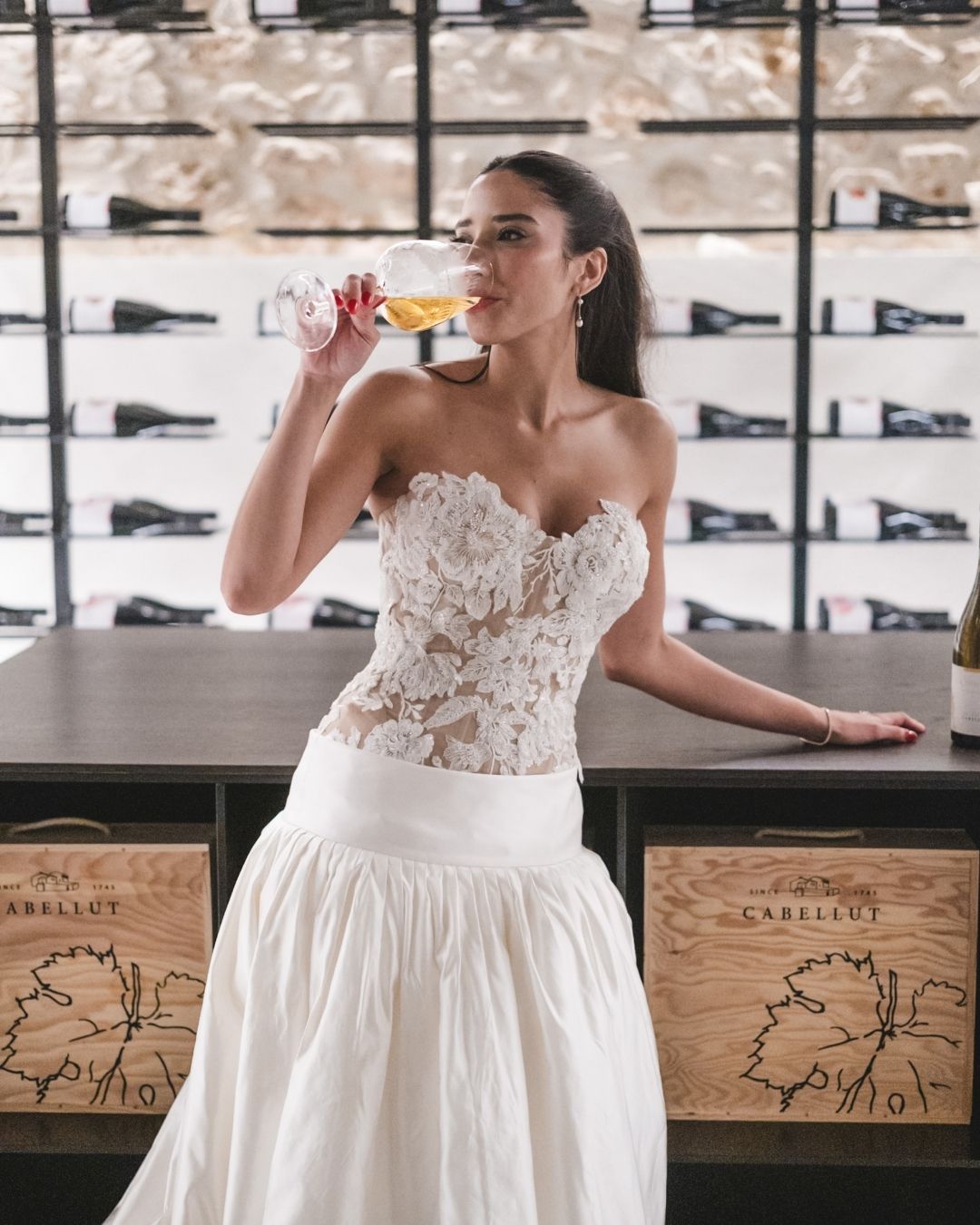 Bride in floral lace gown sipping wine at the Masia Cabellut wine cellar during the wedding celebration.