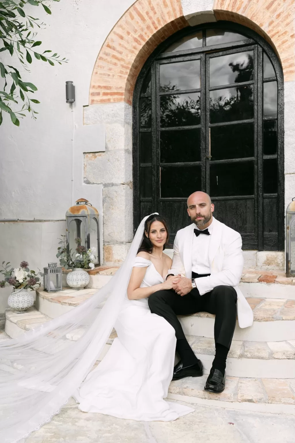 Bride and groom seated on stone steps outside Masia Victoria, holding hands.