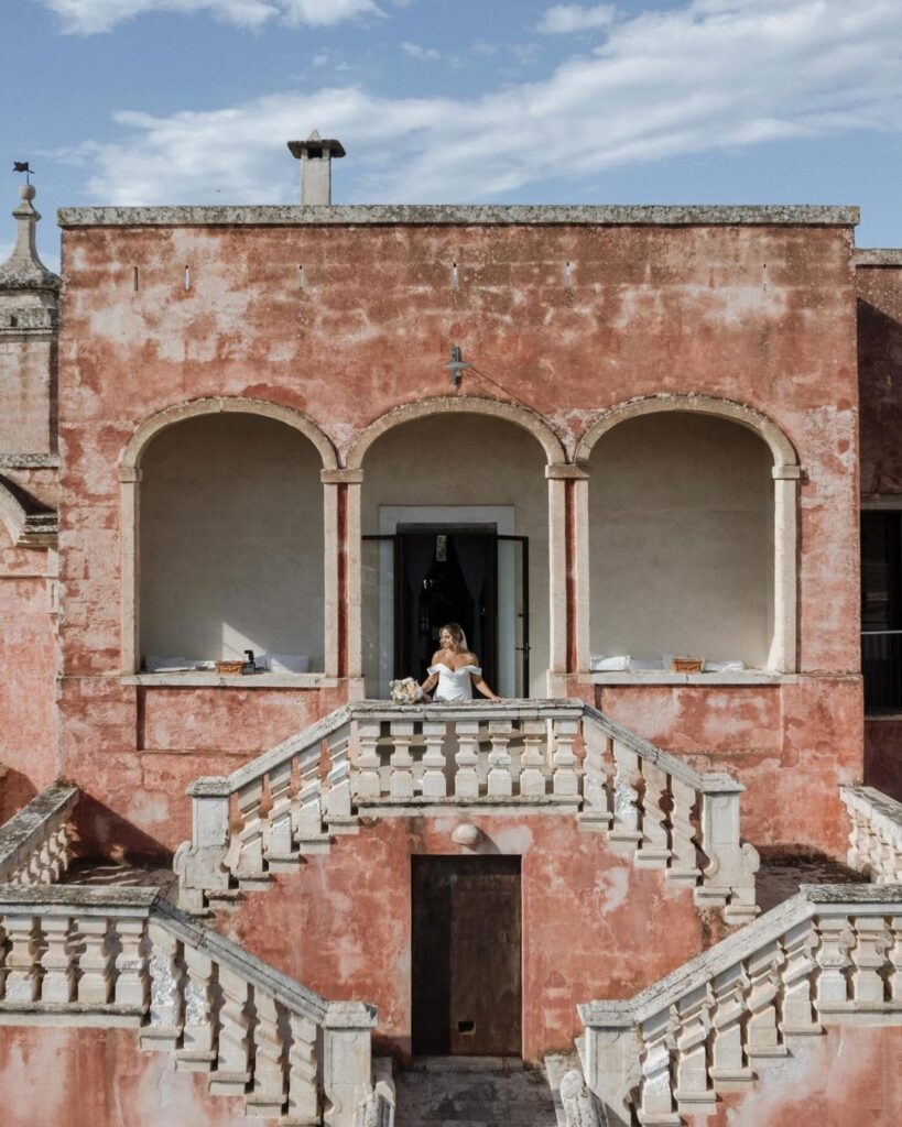 Stunning bride gazing out of a window at Masseria Spina, bathed in natural light and framed by rustic interiors, captured by Amali Studios.