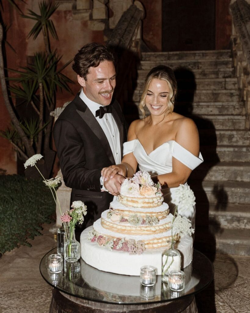 Joyful bride and groom cutting their wedding cake in the courtyard of Masseria Spina, surrounded by candlelight and floral decorations, captured by Amali Studios.