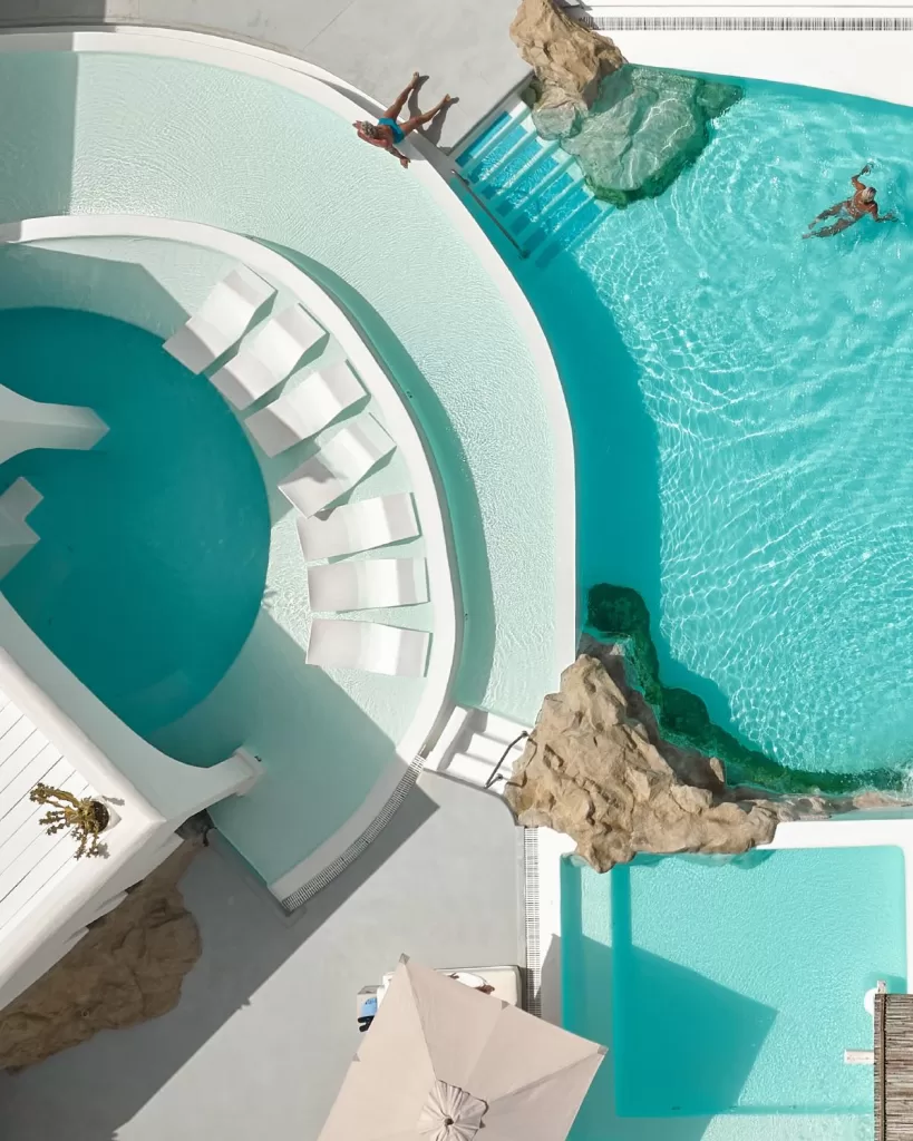 Aerial view of Mykonos Blu’s curved pools with sun loungers and rocky features.