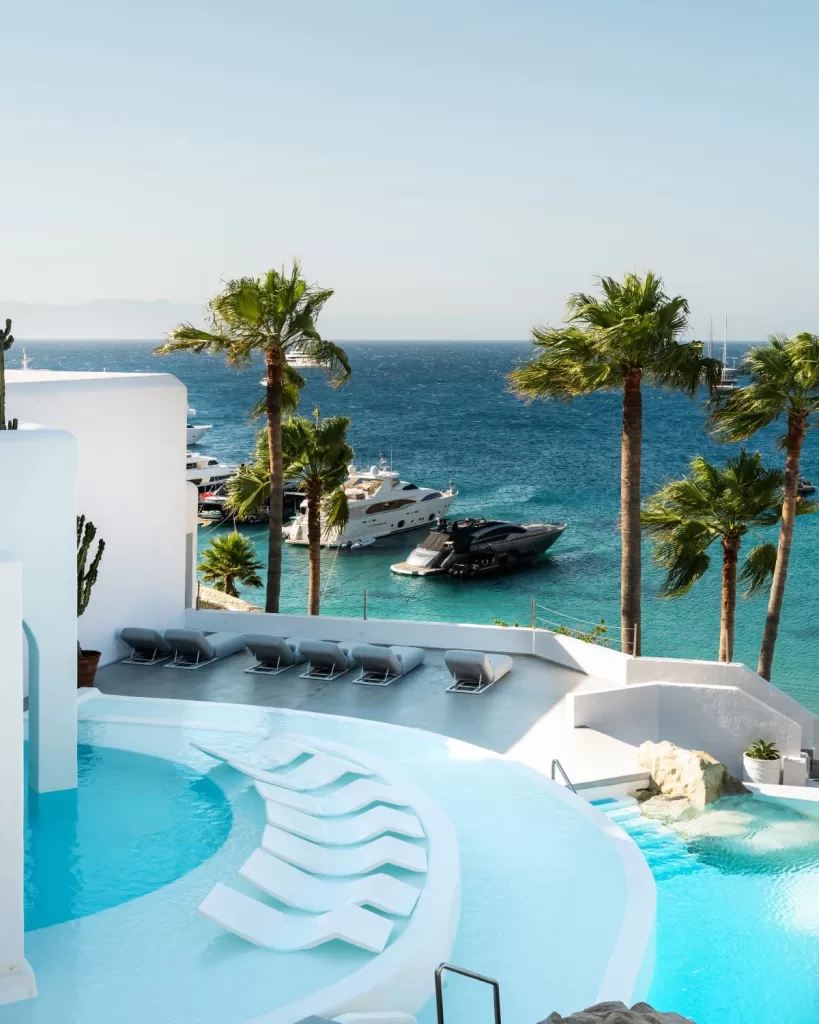 Sun loungers by the curved pool at Mykonos Blu with panoramic views of yachts and the Aegean Sea.