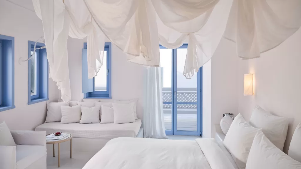 Aegean-inspired suite at Mykonos Blu with floating canopy, soft cushions, and blue-trimmed windows.