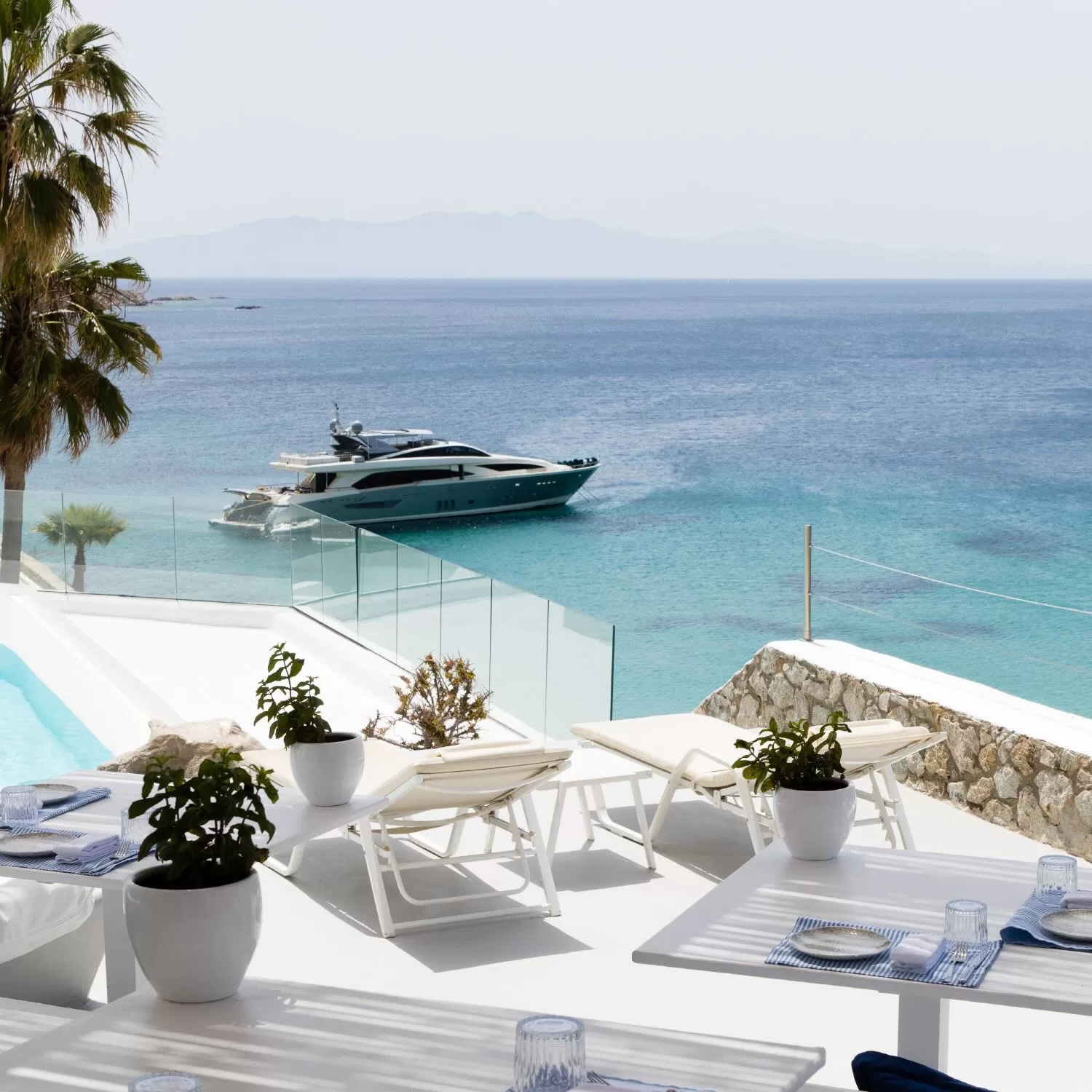 Luxury yacht anchored in the turquoise waters of Psarou Bay, viewed from the chic terrace of Mykonos Blu.