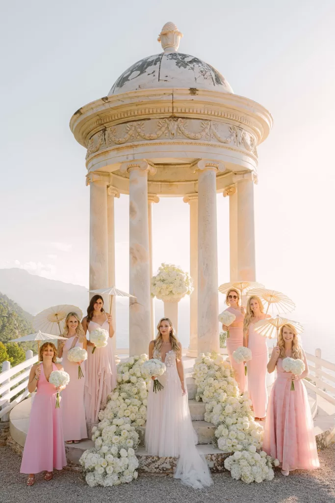 Bride with her bridesmaids in blush dresses holding white bouquets at the Son Marroig pavilion surrounded by romantic floral arrangements.
