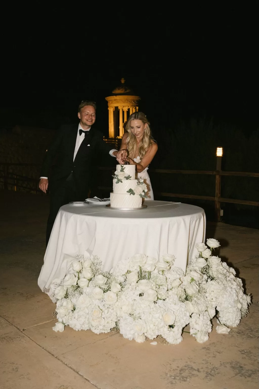 Couple cutting their white wedding cake decorated with roses at Son Marroig, with the illuminated pavilion glowing in the night behind them.