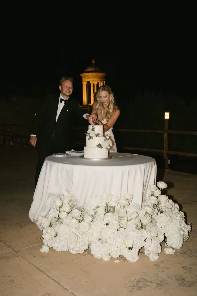Couple cutting their white wedding cake decorated with roses at Son Marroig, with the illuminated pavilion glowing in the night behind them.