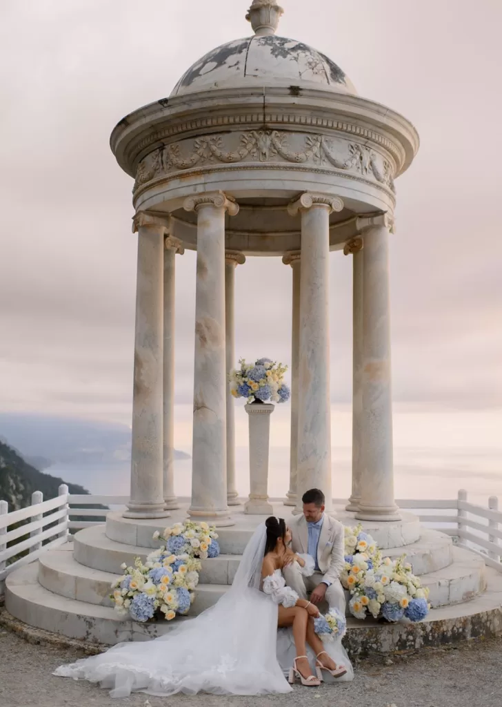 Bride and groom share a tender moment seated on the marble steps of the Son Marroig temple surrounded by blue florals.