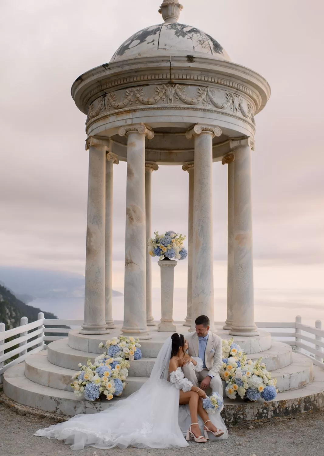 Bride and groom share a tender moment seated on the marble steps of the Son Marroig temple surrounded by blue florals.