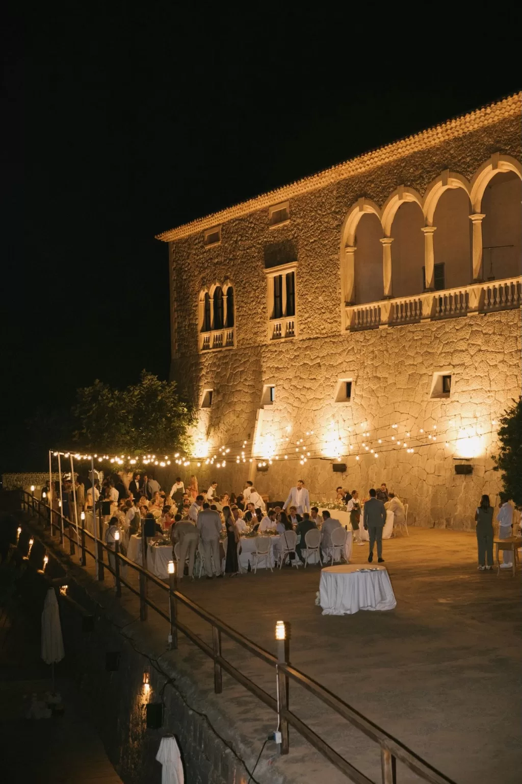 Evening wedding reception at Son Marroig with guests dining under string lights against the stone façade.