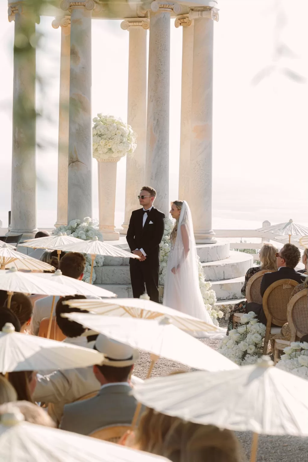 Bride and groom standing beneath the marble pavilion at Son Marroig during their outdoor ceremony, surrounded by guests holding parasols.