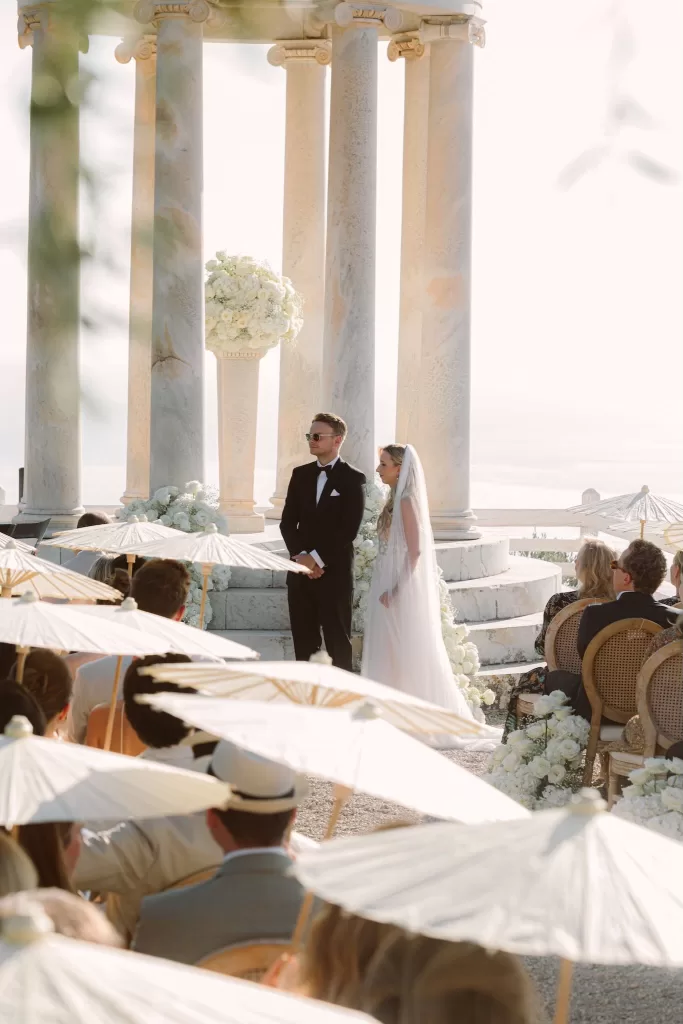 Bride and groom standing beneath the marble pavilion at Son Marroig during their outdoor ceremony, surrounded by guests holding parasols.