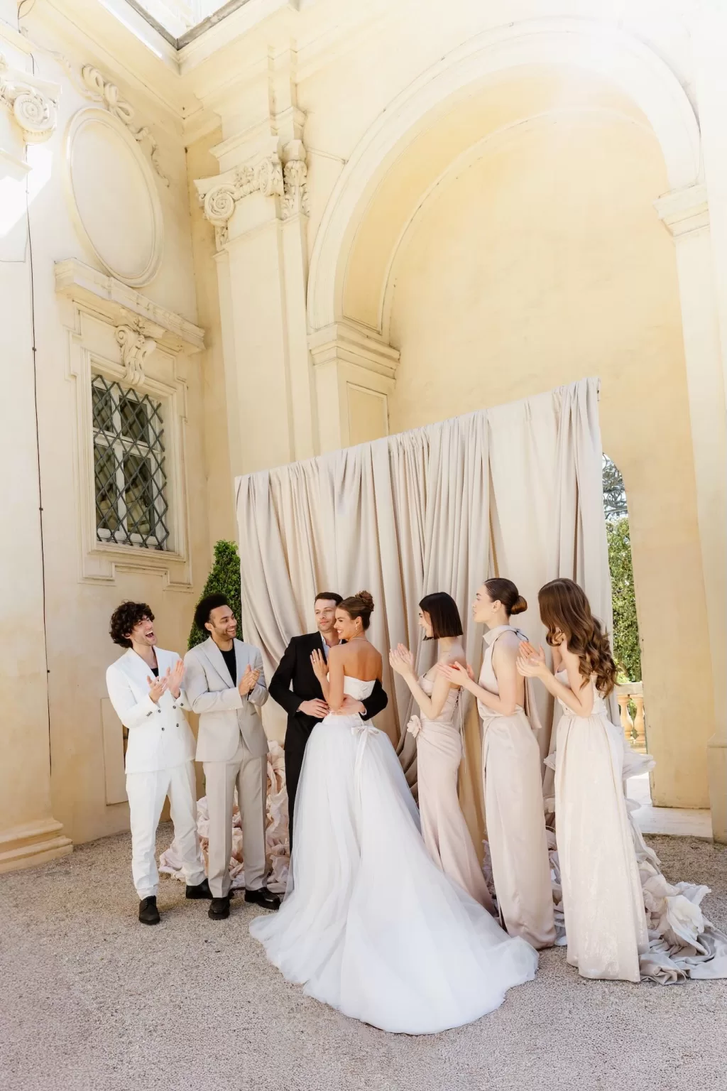 Bride and groom embraced by wedding party in sunlit Villa Aurelia courtyard