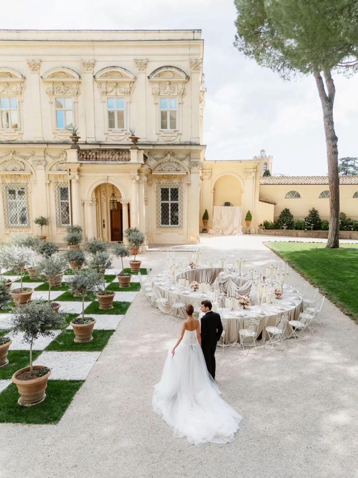 A bride and groom walk toward a circular outdoor reception setup in the courtyard of Villa Aurelia, surrounded by olive trees and historic architecture.