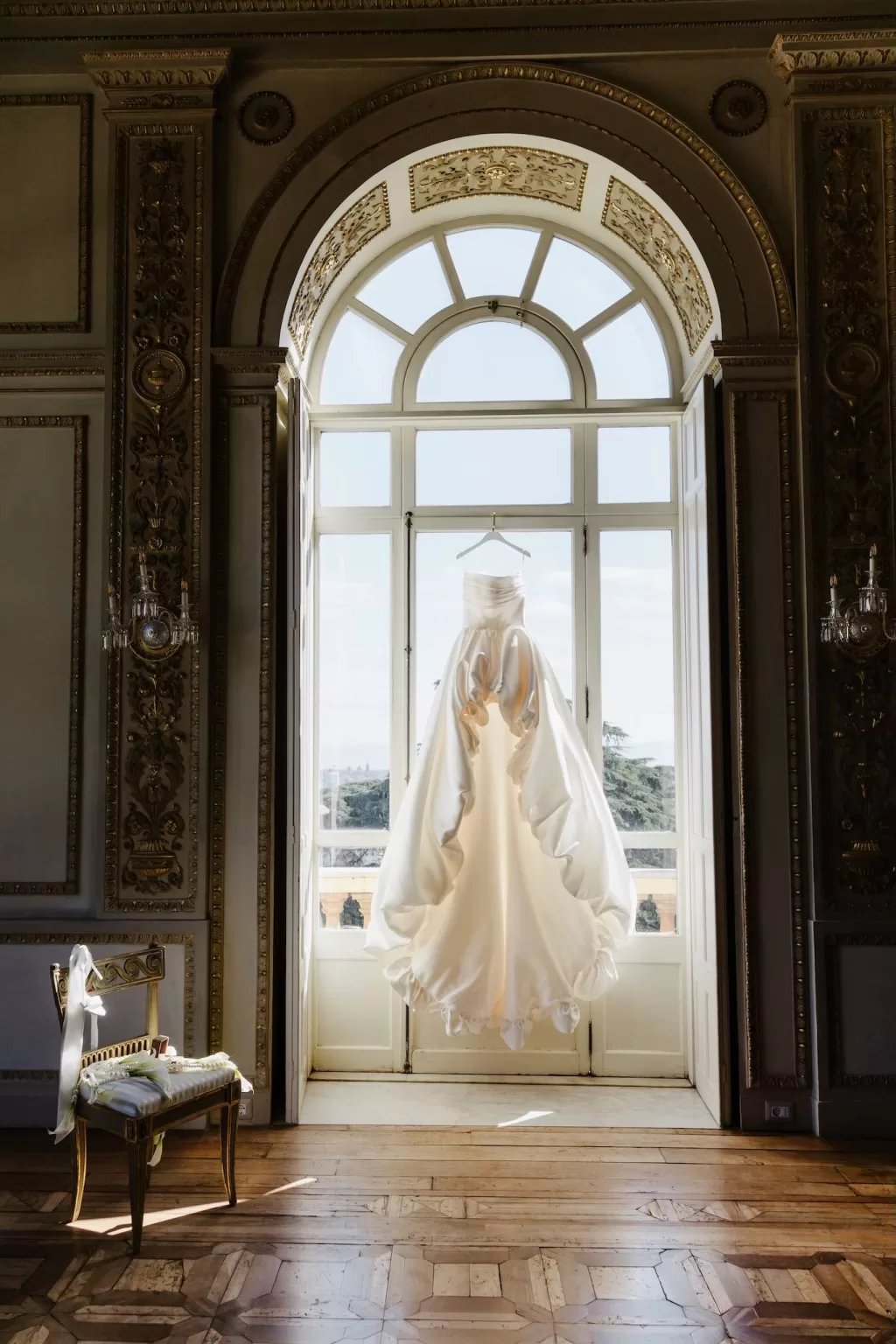 A dramatic wedding gown hangs in the arched window of Villa Aurelia, glowing with morning light.