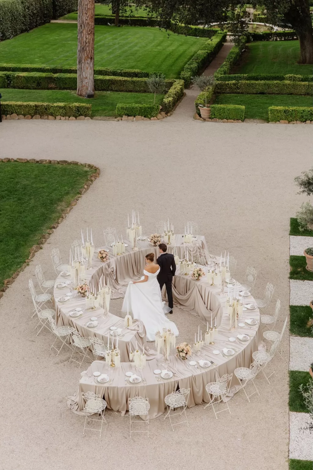 Overhead view of a circular wedding tablescape with neutral linens and candlelight at Villa Aurelia, framed by manicured gardens.