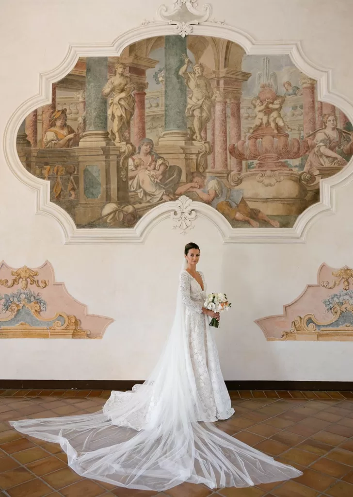 Bride in a lace wedding gown standing beneath an ornate fresco at Villa Cariola, holding a pastel bouquet.