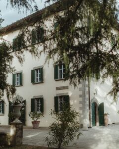 Exterior view of Villa Ivana’s cream facade with green shutters and terracotta pots under a canopy of trees.