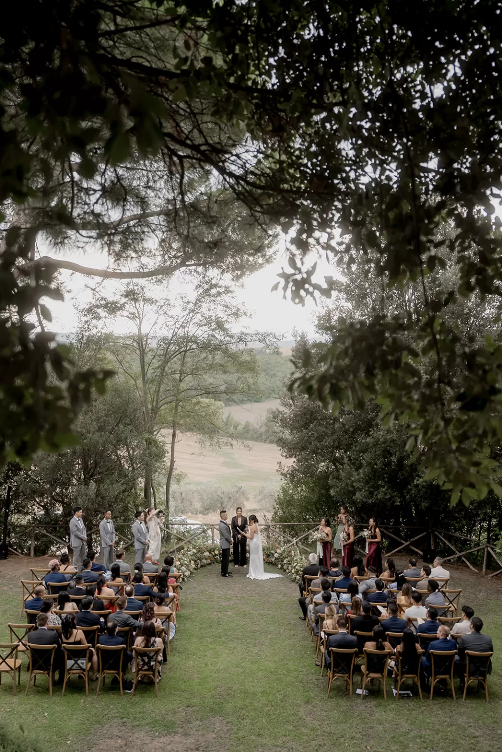 Outdoor ceremony at Borgo Sant Ambrogio framed by trees, with guests seated beneath the Tuscan sky and rolling hills beyond.