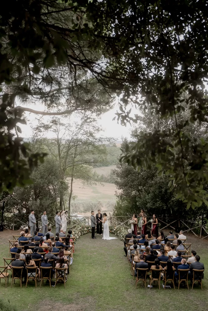 Outdoor ceremony at Borgo Sant Ambrogio framed by trees, with guests seated beneath the Tuscan sky and rolling hills beyond.