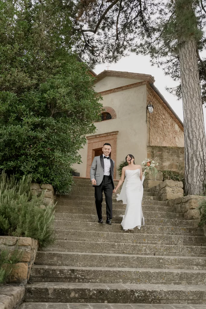 Newlyweds descending the stone steps in front of the chapel at Borgo Sant Ambrogio, hand in hand beneath tall cypress trees.