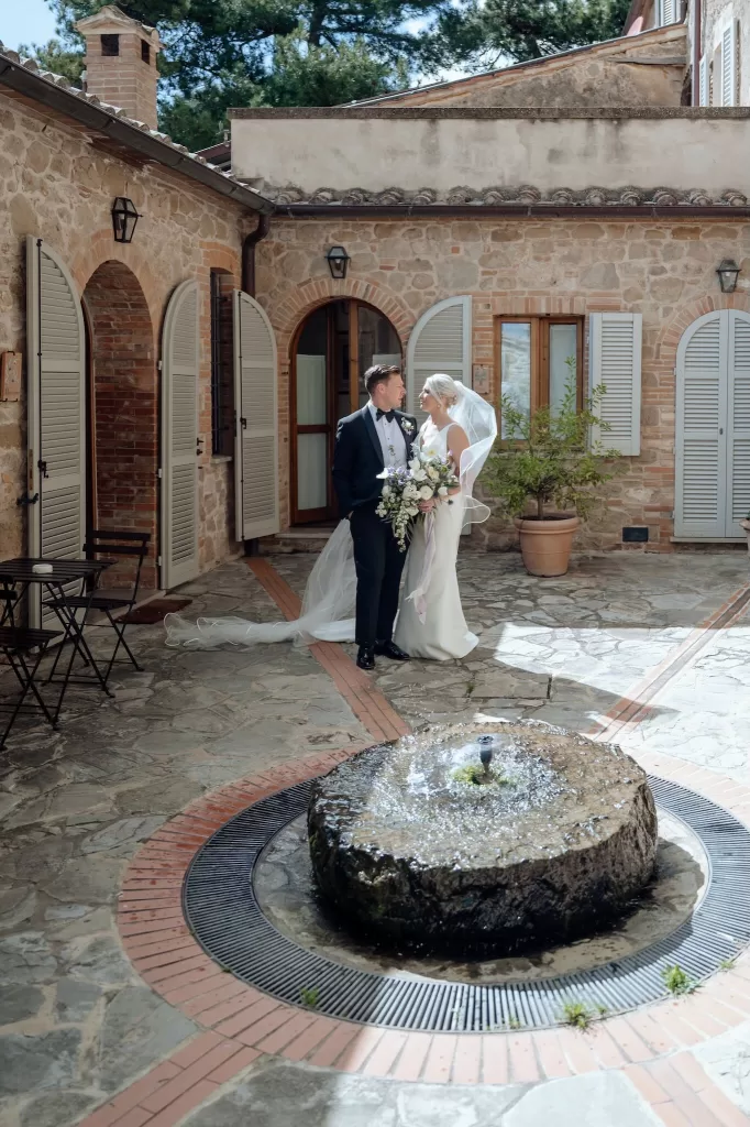 Newlyweds pose by the courtyard fountain surrounded by rustic stone walls at Borgo Sant'Ambrogio.