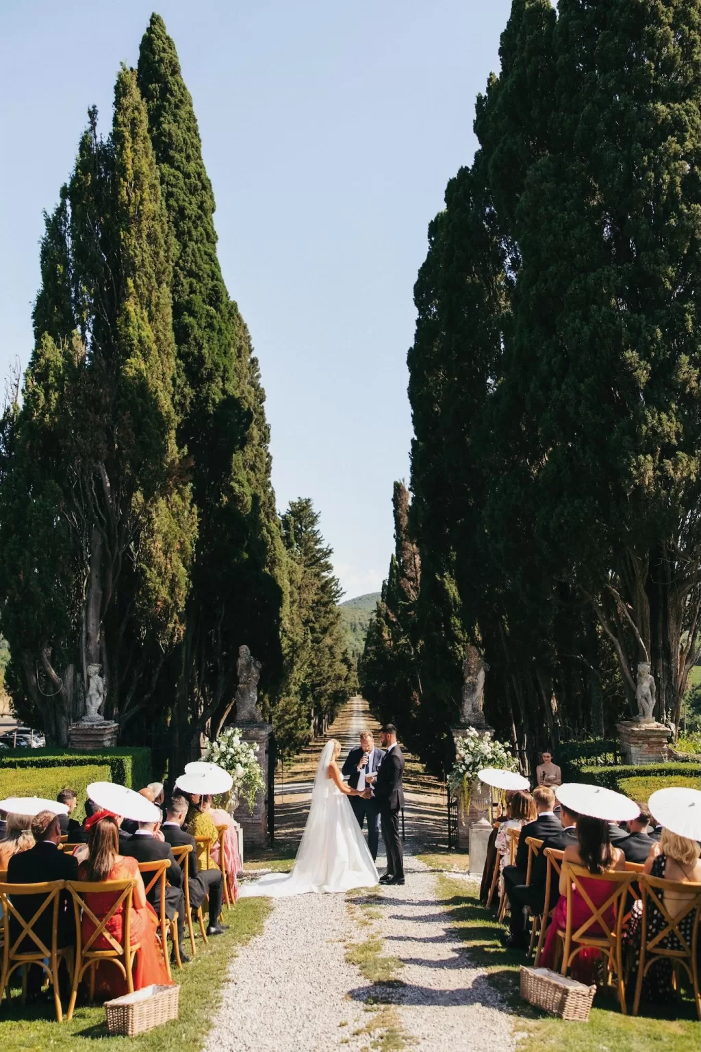 Outdoor ceremony under tall cypress trees at Borgo Stomenanno with guests seated on either side.