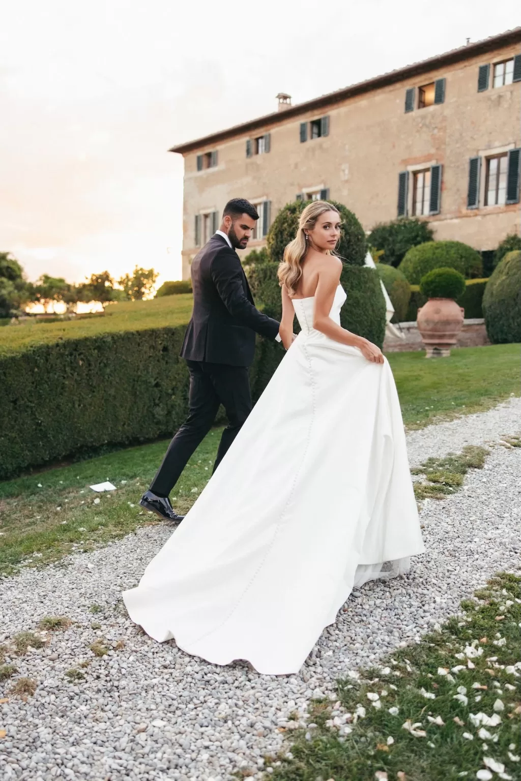 Bride and groom walking through the garden at sunset, with Borgo Stomenanno’s villa in the background.