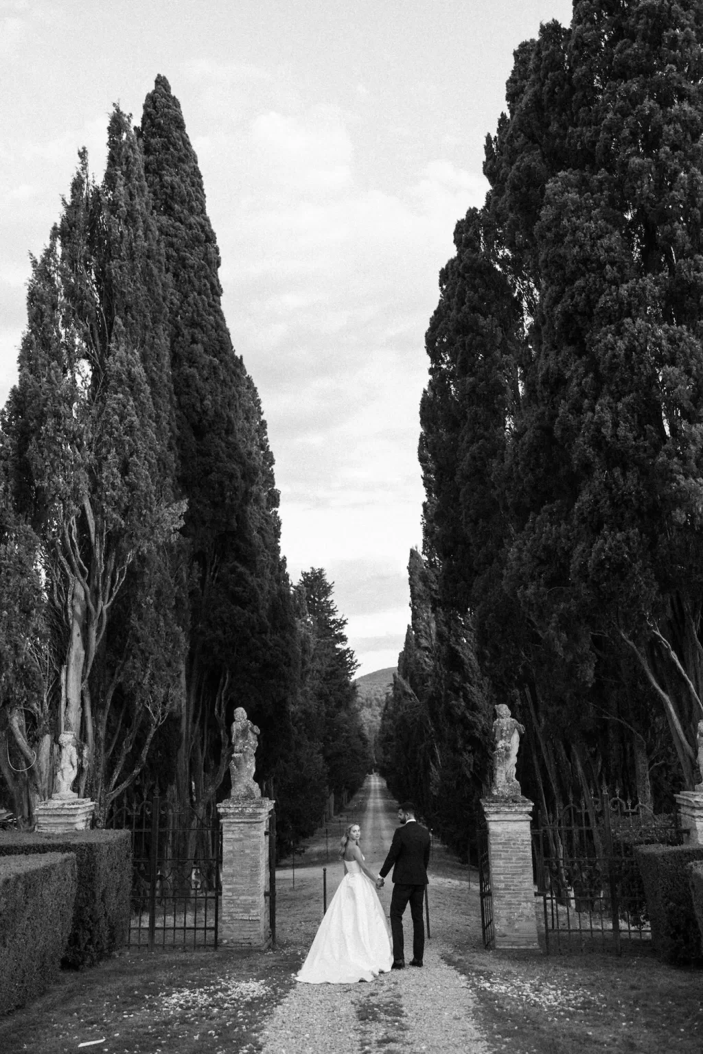 The newlyweds walk hand in hand down the cypress-lined avenue at Borgo Stomenanno.
