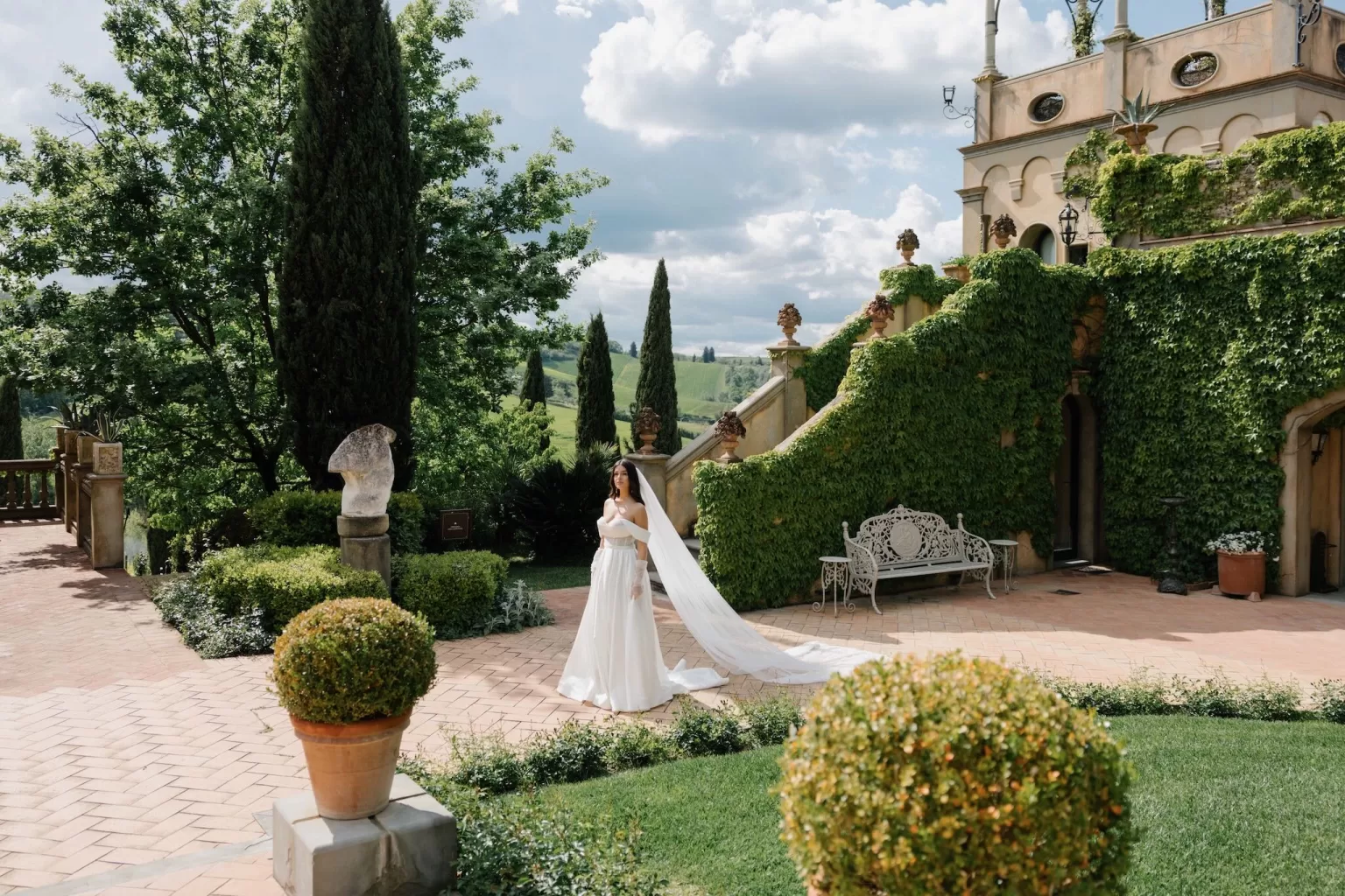 A bride in an elegant gown with a long veil walking through the ivy-covered courtyard of Tenuta Corbinaia, surrounded by Tuscan cypress trees.