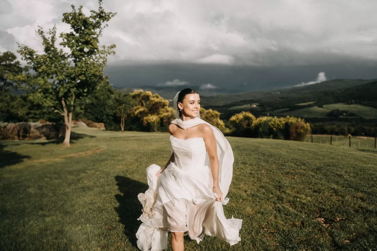 Bride runs through sunlit grass at Castello di Casole, with a sweeping Tuscan landscape behind her.