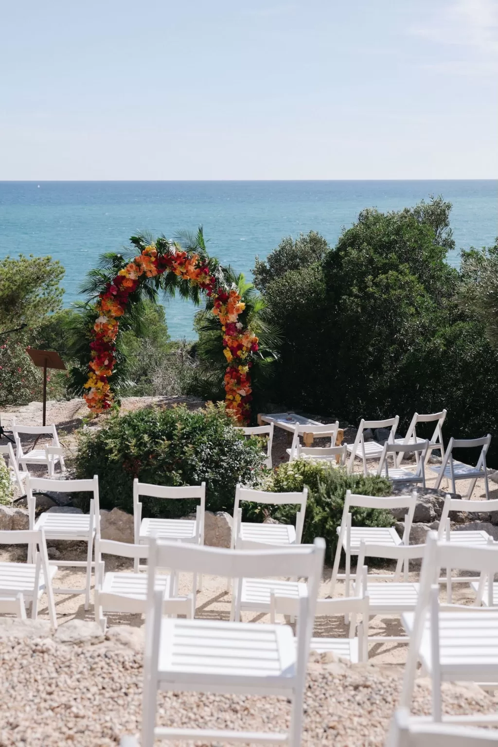 Vibrant floral arch and white chairs arranged for a seaside ceremony at Masia Casa del Mar.
