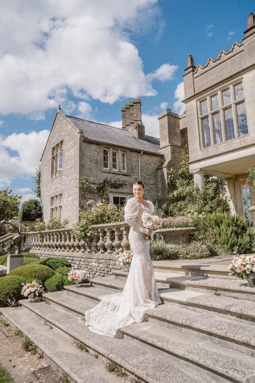 The bride poses on stone steps in front of the grand Euridge Manor, holding a blush-toned bouquet under sunny skies.