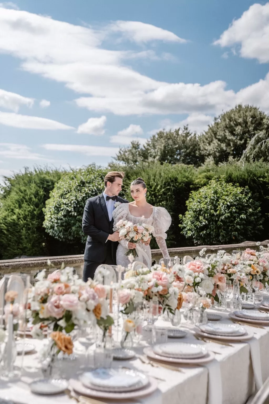 Bride and groom pose by a floral-filled wedding reception table under blue skies at Euridge Manor.