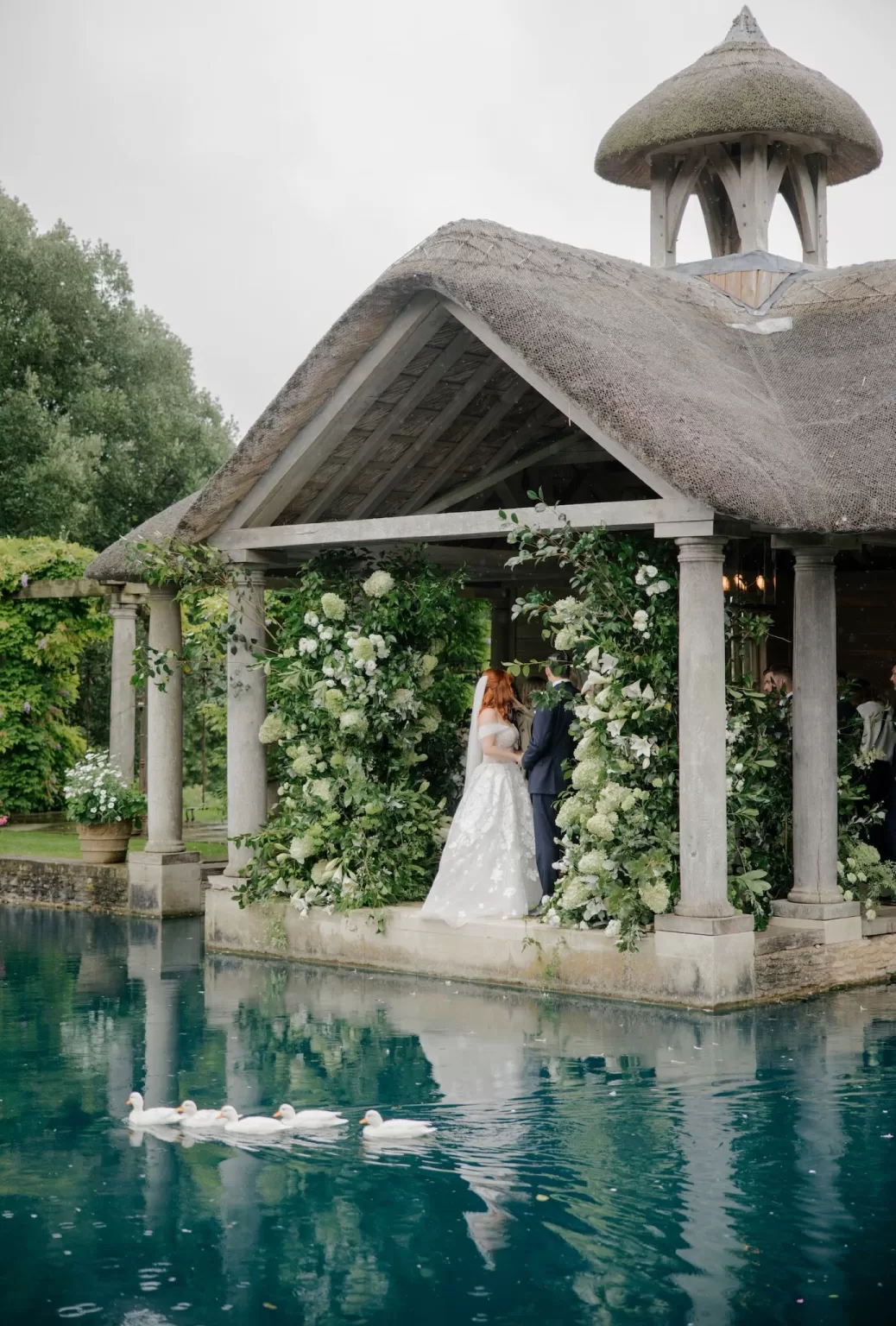 The couple exchanges vows beneath the pavilion at Euridge Manor, while a line of ducks glides across the water.