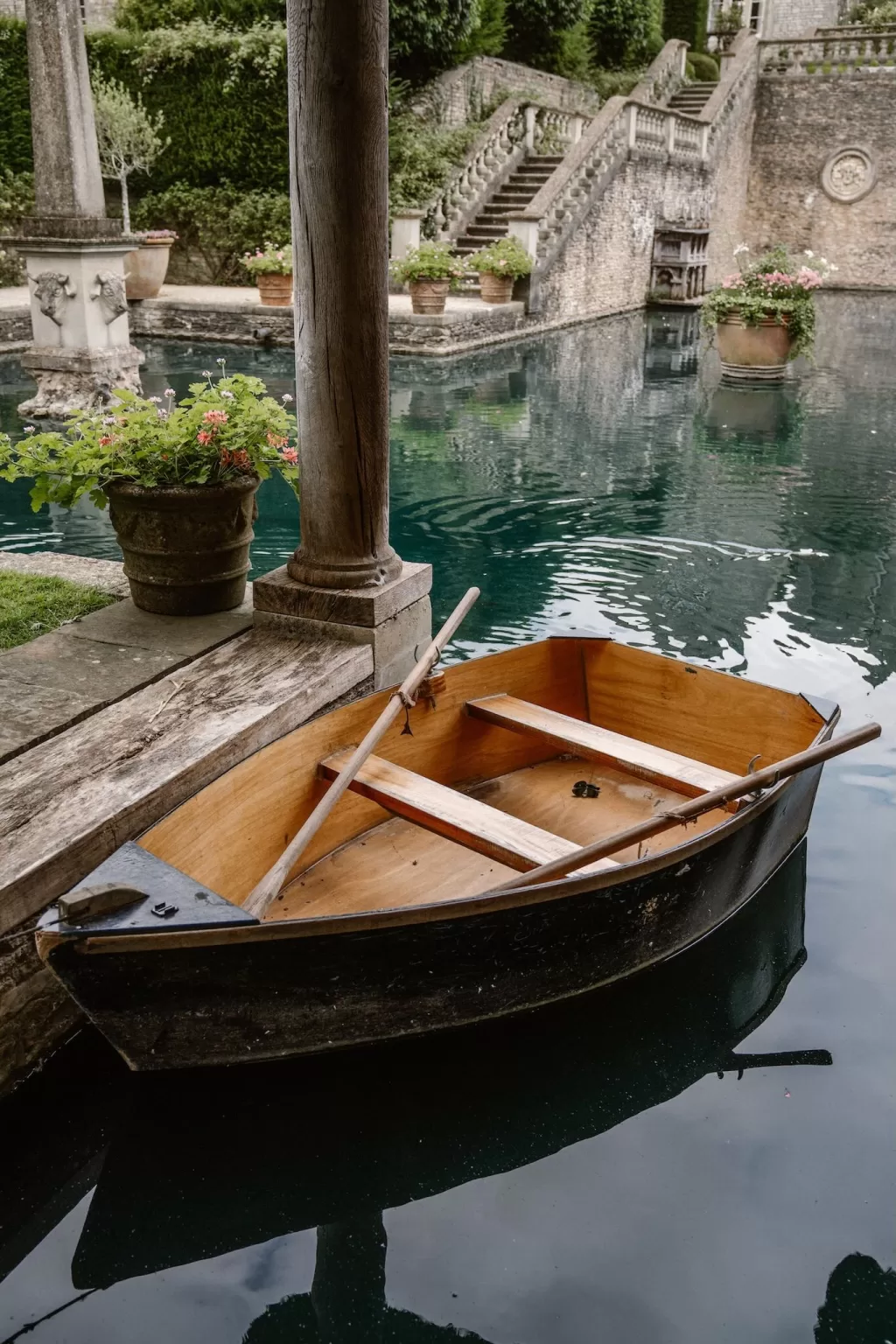 A wooden rowboat floats gently on the still turquoise pond at Euridge Manor, surrounded by stone columns and lush greenery.