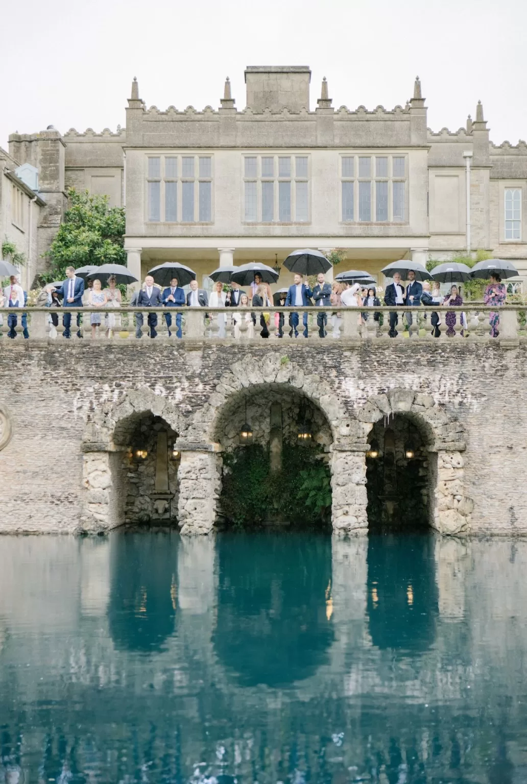 Wedding guests holding umbrellas on the stone terrace at Euridge Manor, overlooking the turquoise water below.