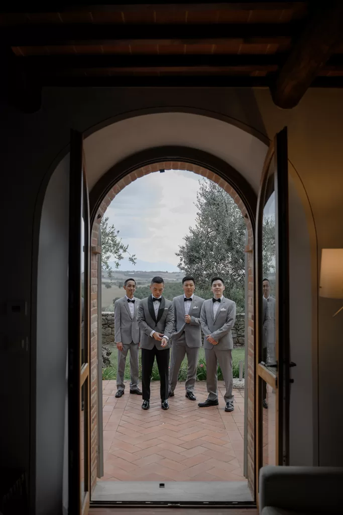 Groomsmen line up outside arched wooden door with countryside views behind them at Borgo Sant'Ambrogio.