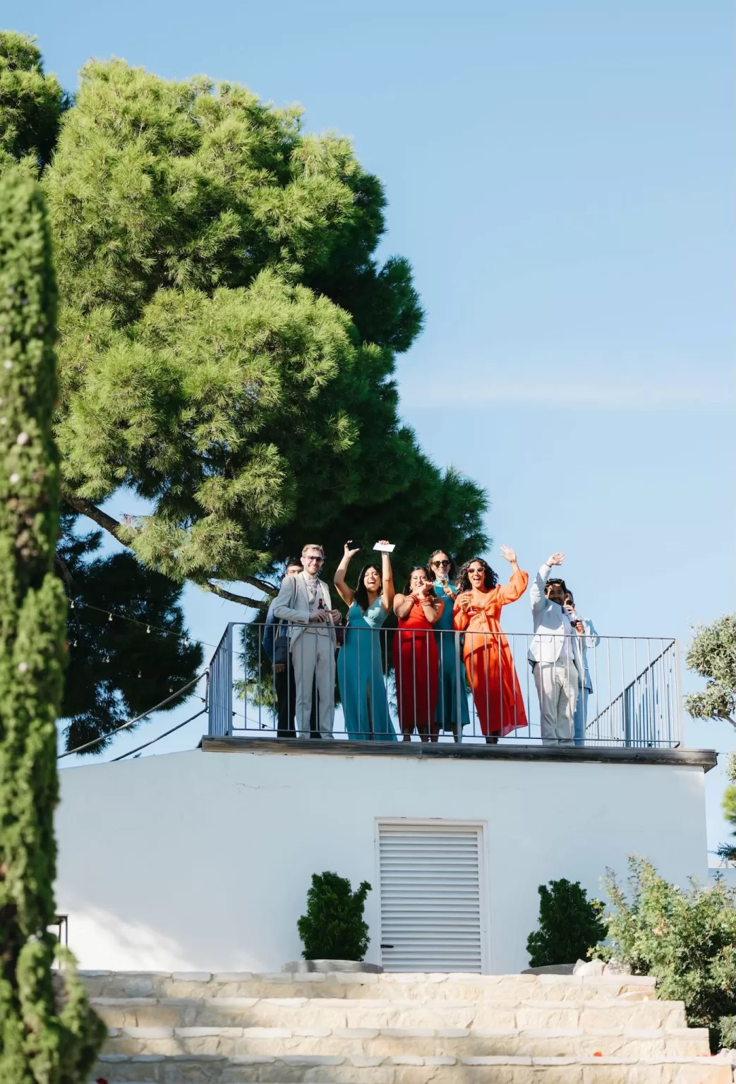 Wedding guests waving from a balcony surrounded by pine trees and clear skies at Masia Casa del Mar.