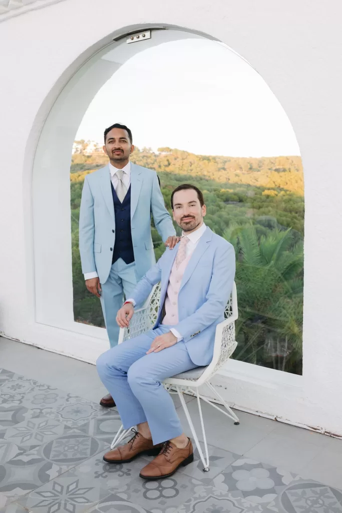 Wedding portrait of two grooms in powder blue suits on the tiled balcony of Masia Casa del Mar, framed by lush hillside views.