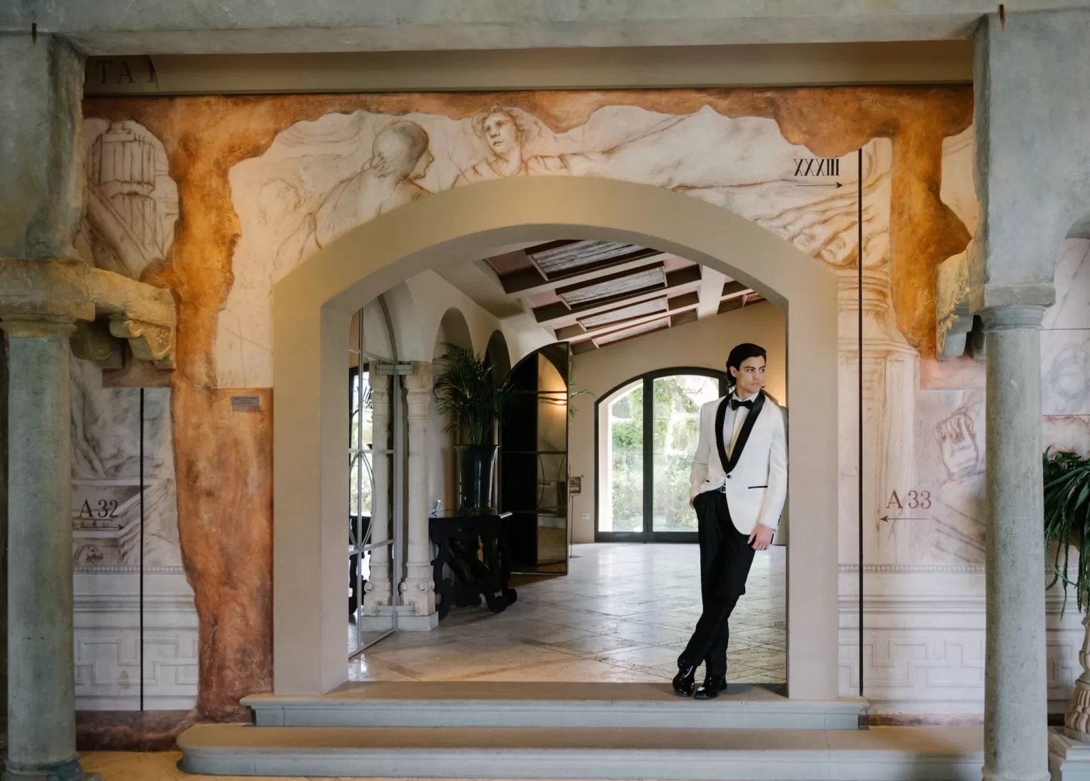 Groom in a white tuxedo standing in the frescoed archway of Tenuta Corbinaia, blending modern style with Italian heritage.