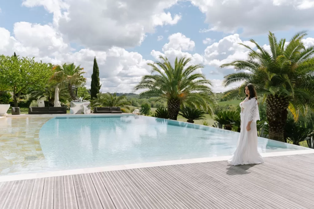Bride in a white robe walking beside the palm-fringed pool at Tenuta Corbinaia with rolling Tuscan hills in the distance.