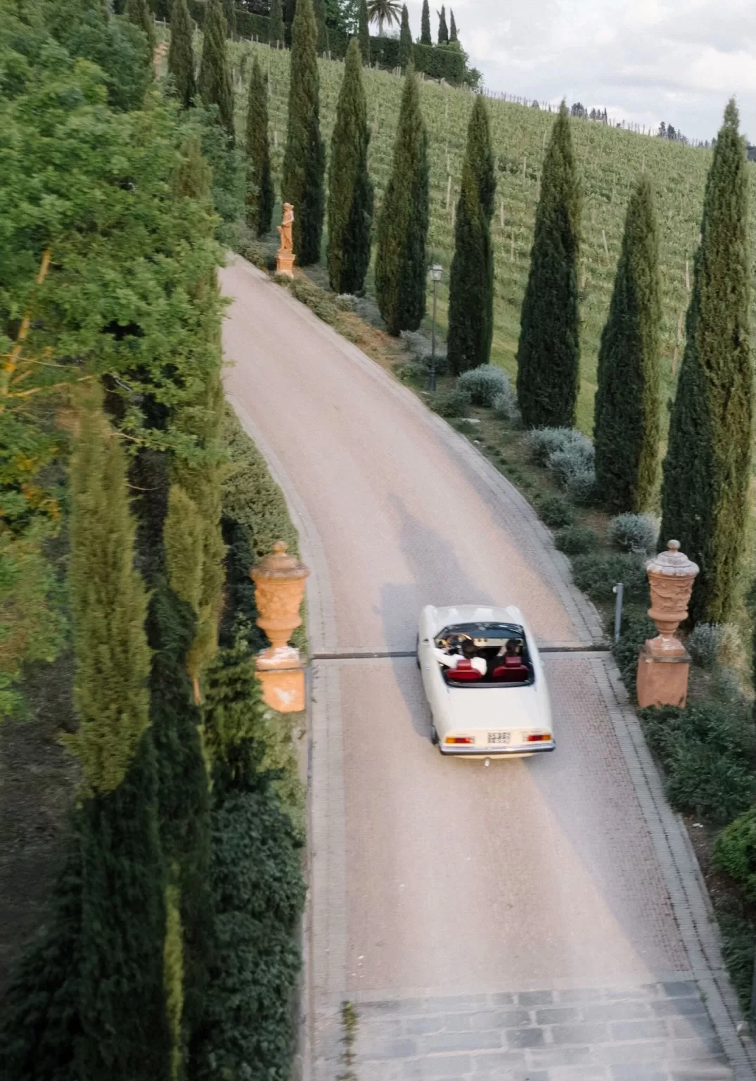 Couple driving a vintage convertible up the cypress-lined driveway of Tenuta Corbinaia, setting a romantic Tuscan arrival scene.