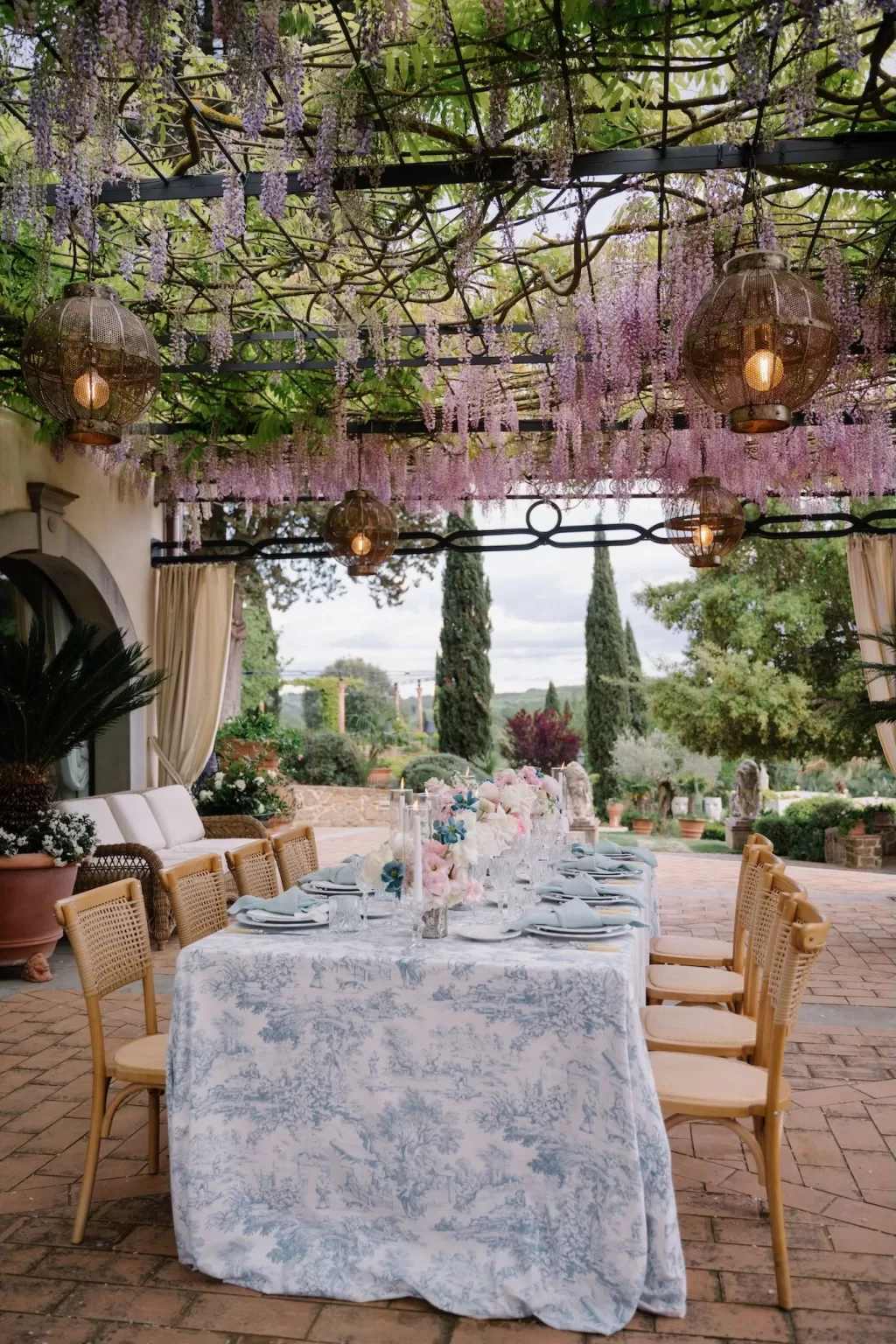 Long reception table under blooming wisteria pergola at Tenuta Corbinaia with views of Tuscan gardens.